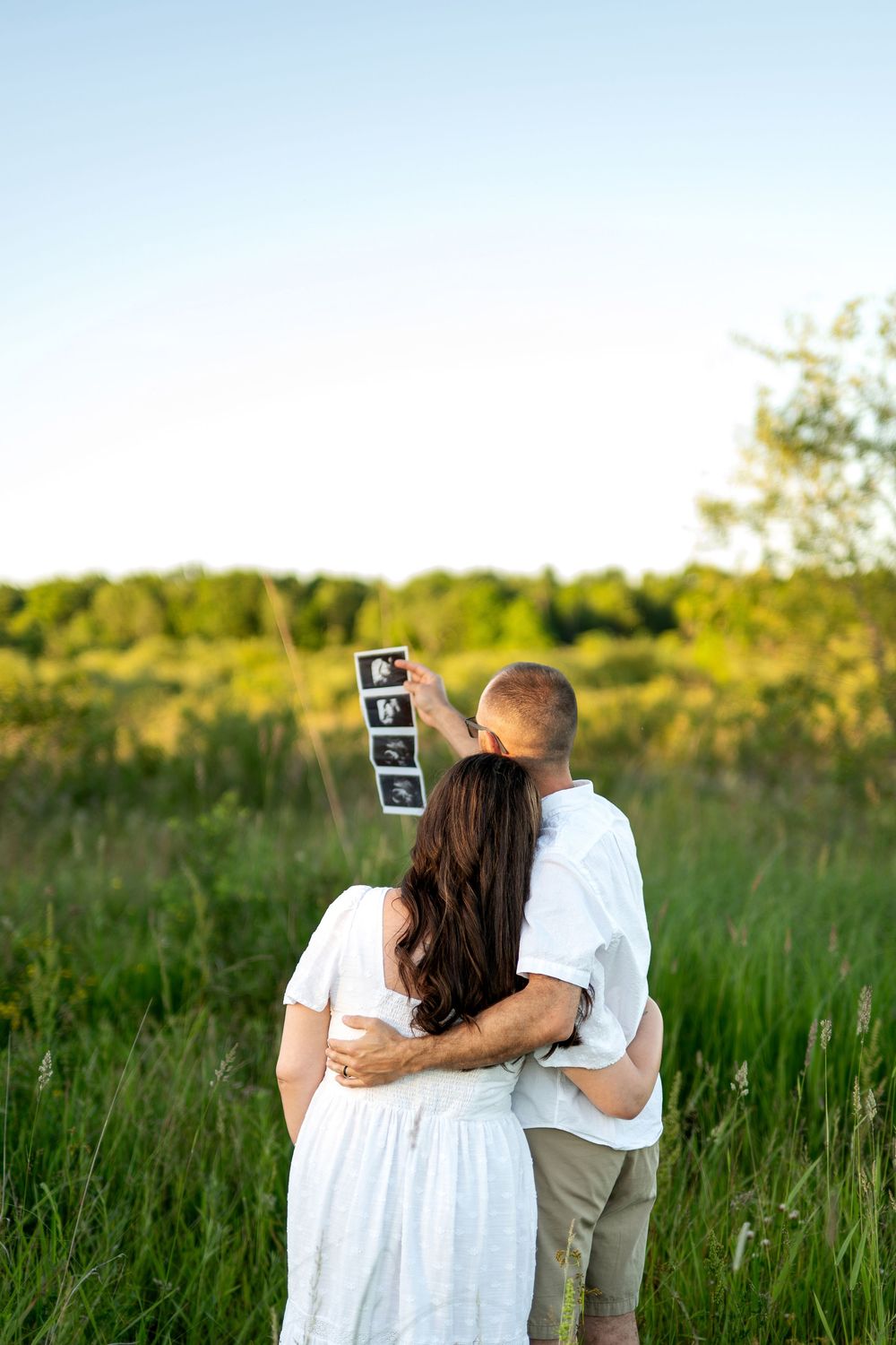 Couple standing in green field holding ultrasound photos, woman in white dress with long dark hair, man in white shirt embracing her.