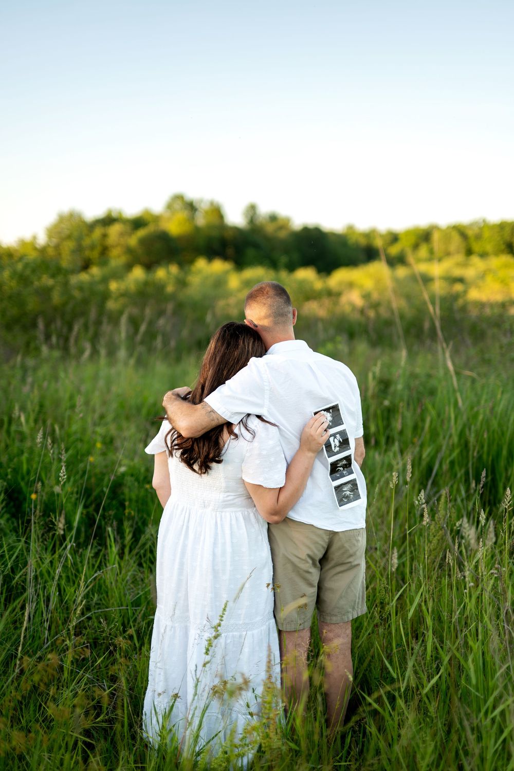 Couple embracing from behind in a green meadow, man holding ultrasound strip photos on his back.