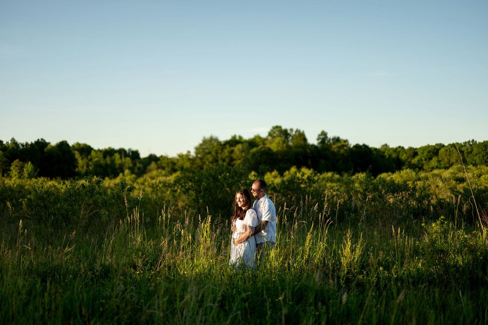 Expecting couple standing together in a sunlit green meadow at golden hour, surrounded by tall grass.