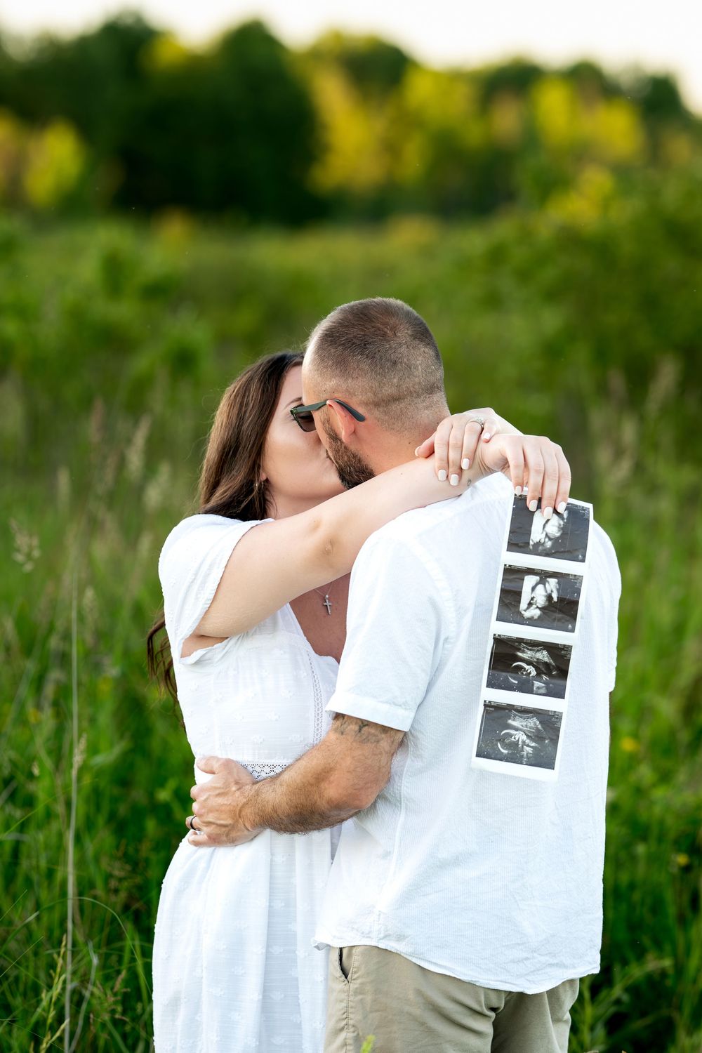 Couple kissing while holding ultrasound images in a green meadow during golden hour.