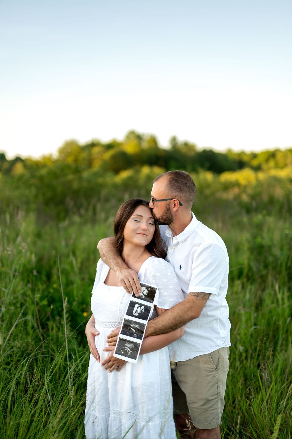 Man kissing pregnant woman's temple while she holds ultrasound photos in a sunny green field.