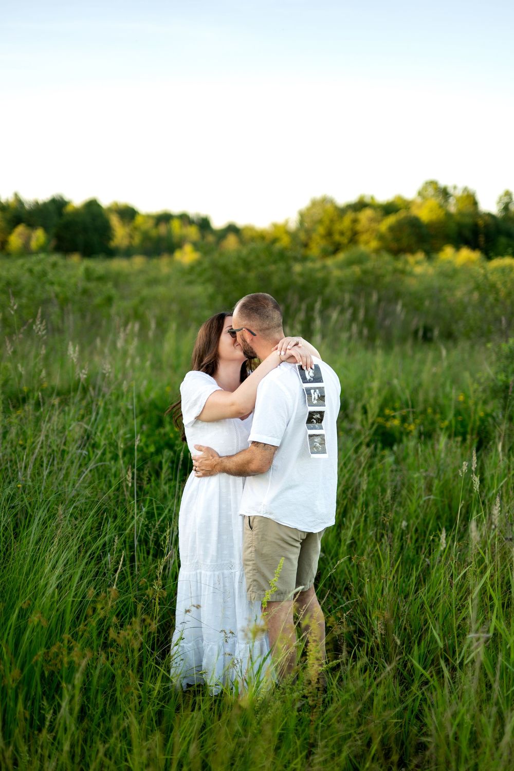 Couple kissing in a lush green field, woman in white dress embracing man in white t-shirt outdoors.