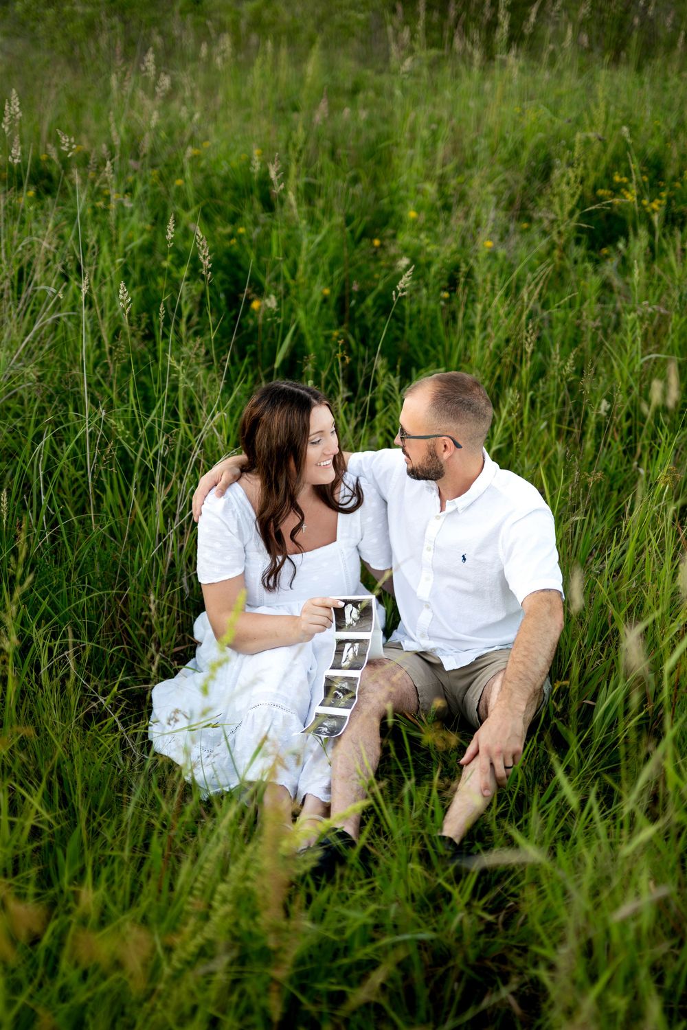Pregnant couple sitting in tall green grass, smiling at each other while holding ultrasound images.
