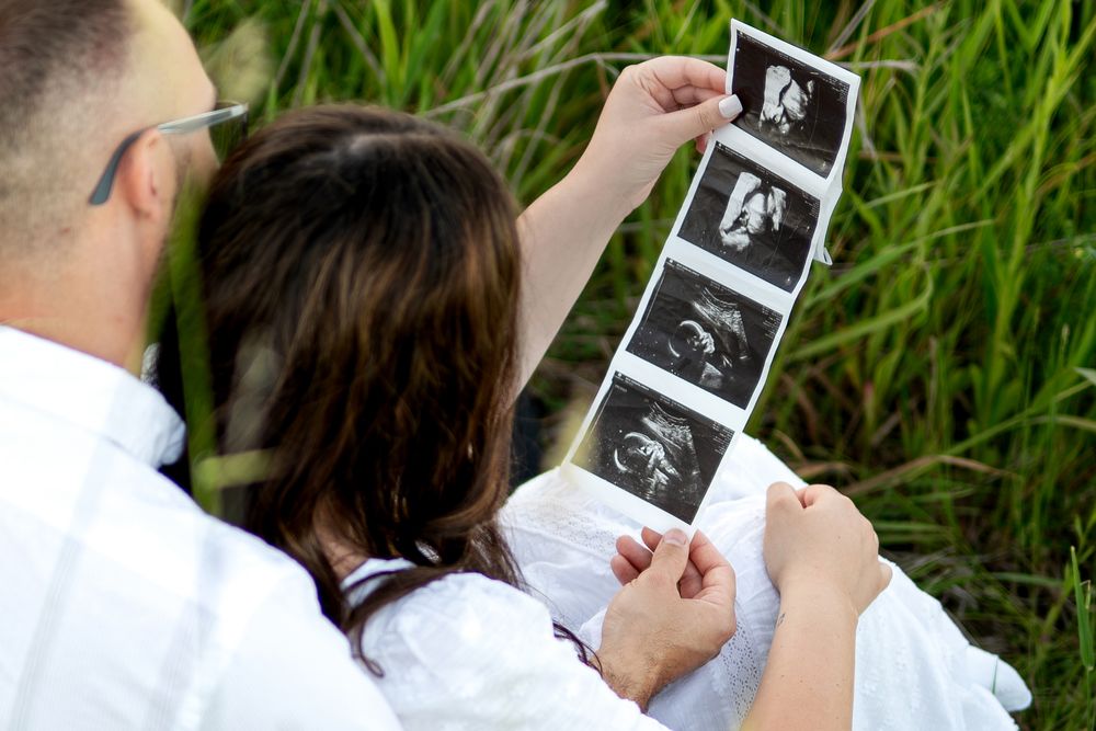 Expecting couple sitting in grass reviewing baby ultrasound photos together outdoors.