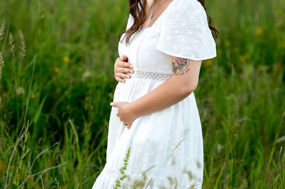 Pregnant woman in white dress holding baby bump in green field.