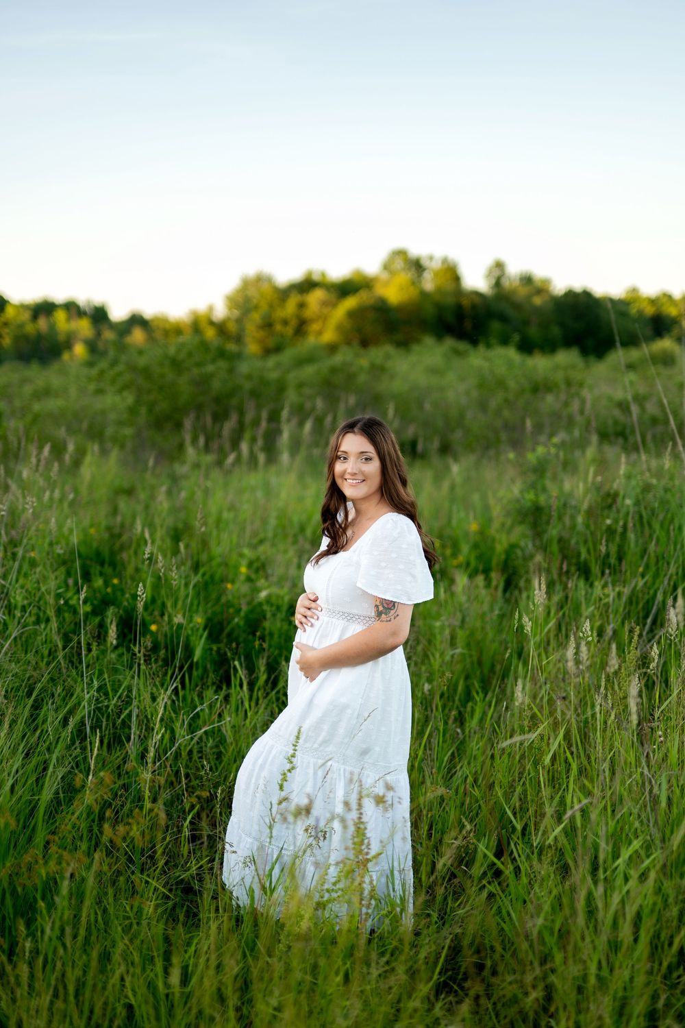 Smiling pregnant woman in white dress cradling baby bump in lush green field at sunset.