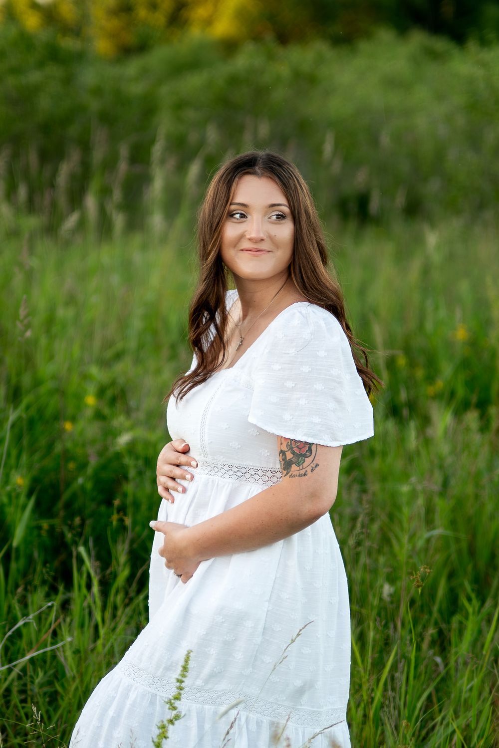 Pregnant woman in white dress standing in green field, cradling baby bump, with tattoo on arm, smiling outdoors.