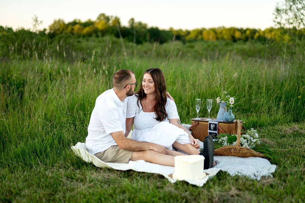 Couple enjoying a romantic outdoor picnic on a white blanket in a green field with wine glasses and a basket.