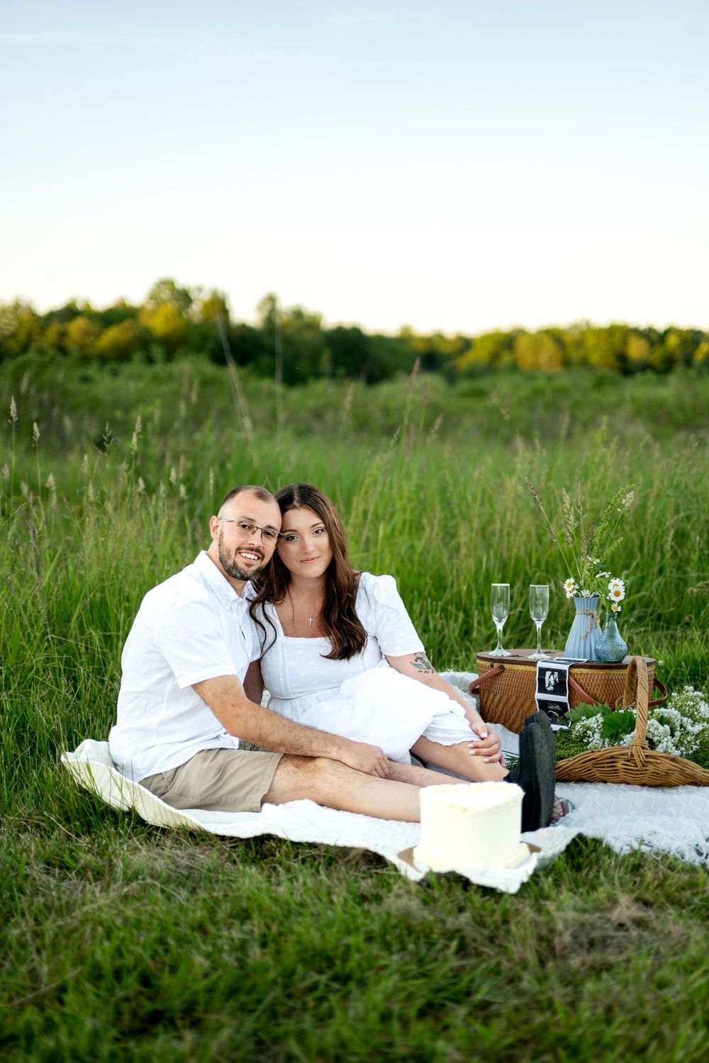 Couple sitting on white blanket enjoying a romantic picnic in a green meadow with champagne glasses.
