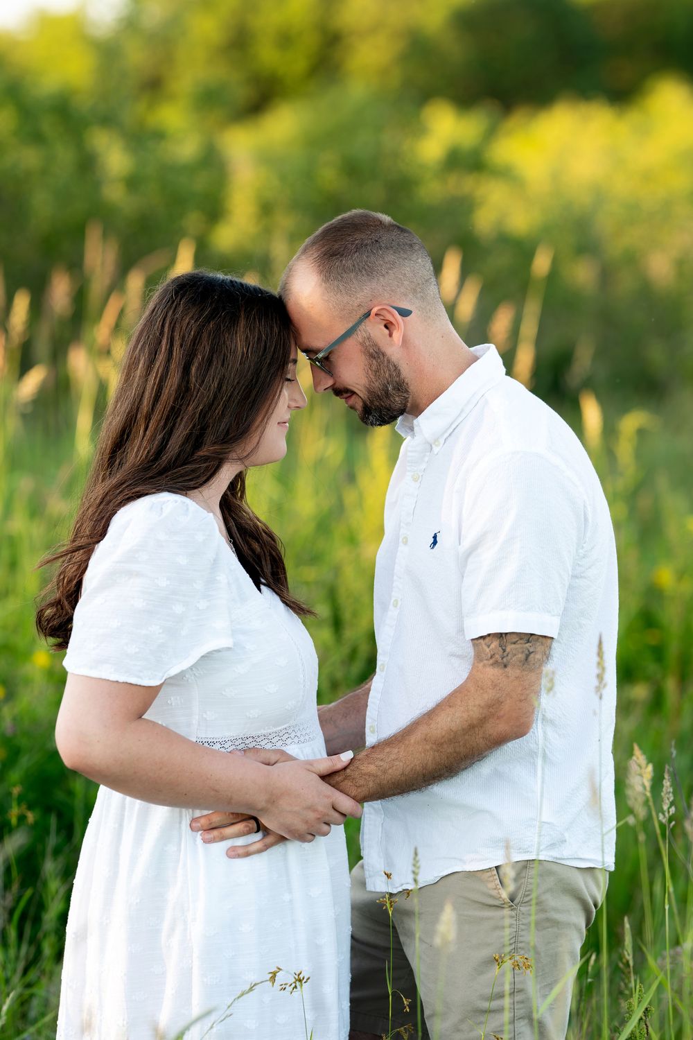 Couple tenderly touching foreheads in a lush green field, man holding pregnant woman's belly in white dress.