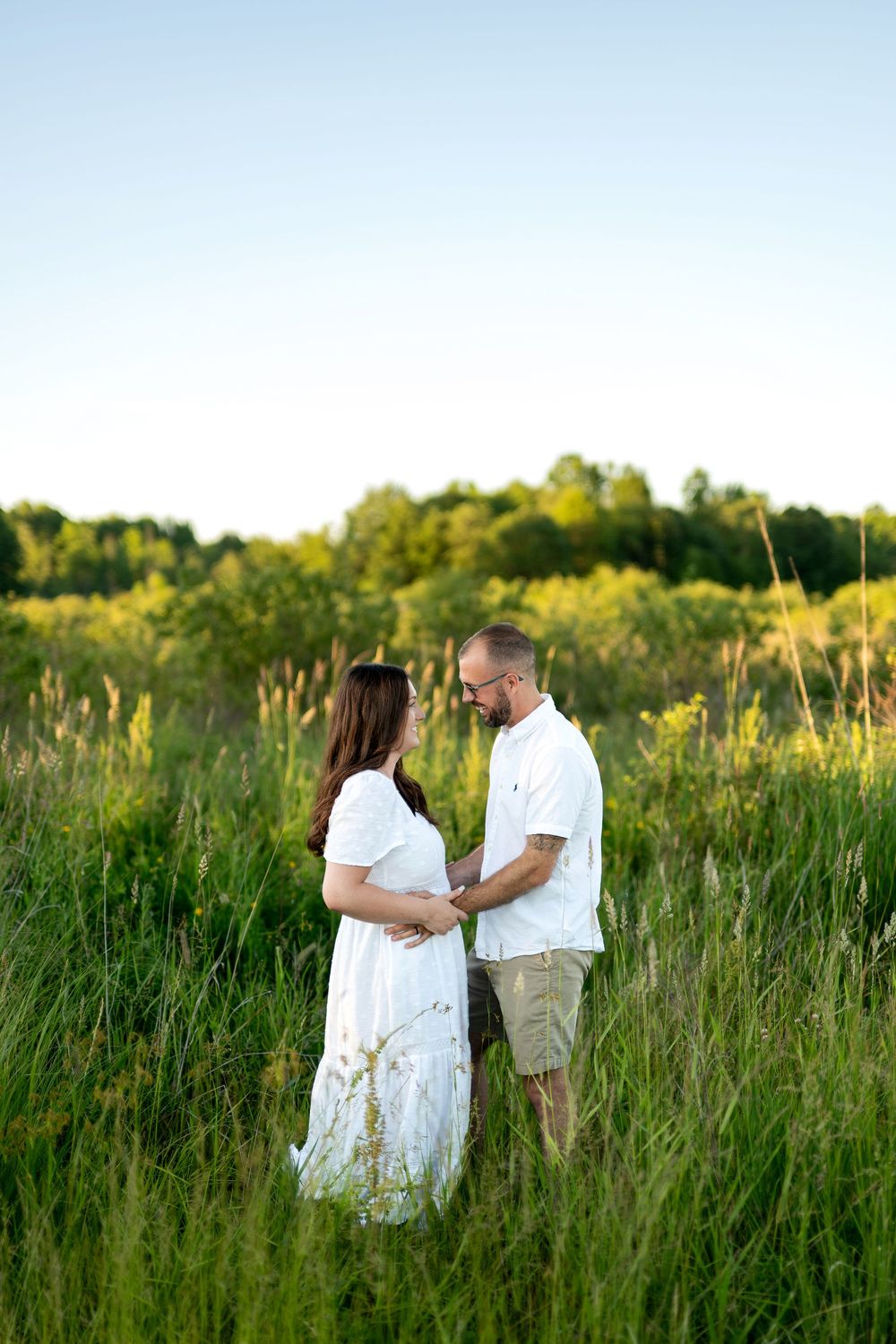 Couple holding hands in a lush green field, woman in white dress facing man in white shirt and khaki shorts.