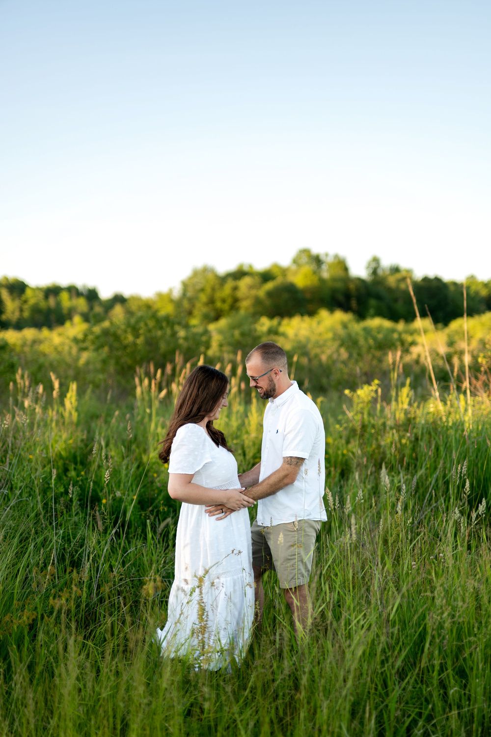 Pregnant couple holding hands in a lush green field at golden hour, woman in white dress.