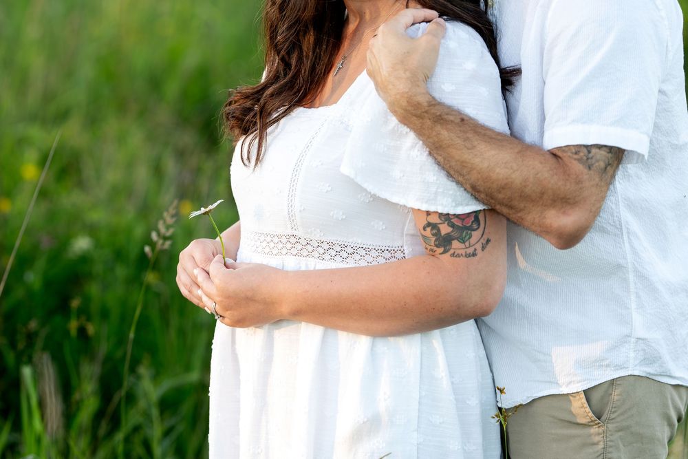 Couple embracing outdoors in green field, woman in white dress, man with colorful tattoo on arm.