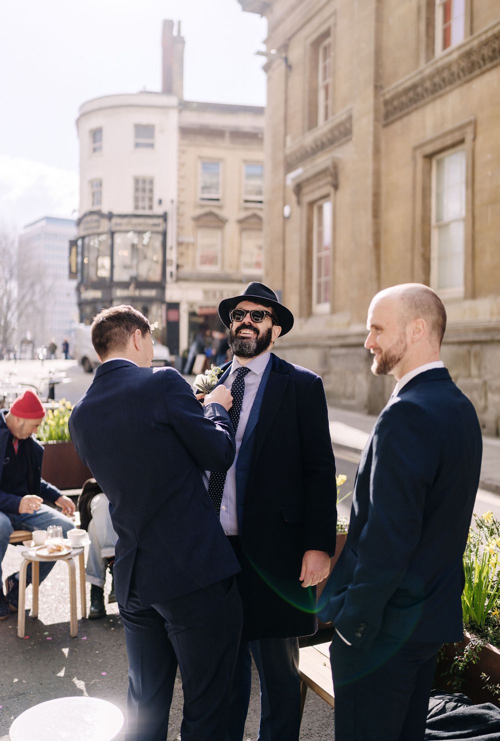 Three groomsmen in navy suits chat outside a historic building on a sunny wedding day in Bristol, corn street registry office