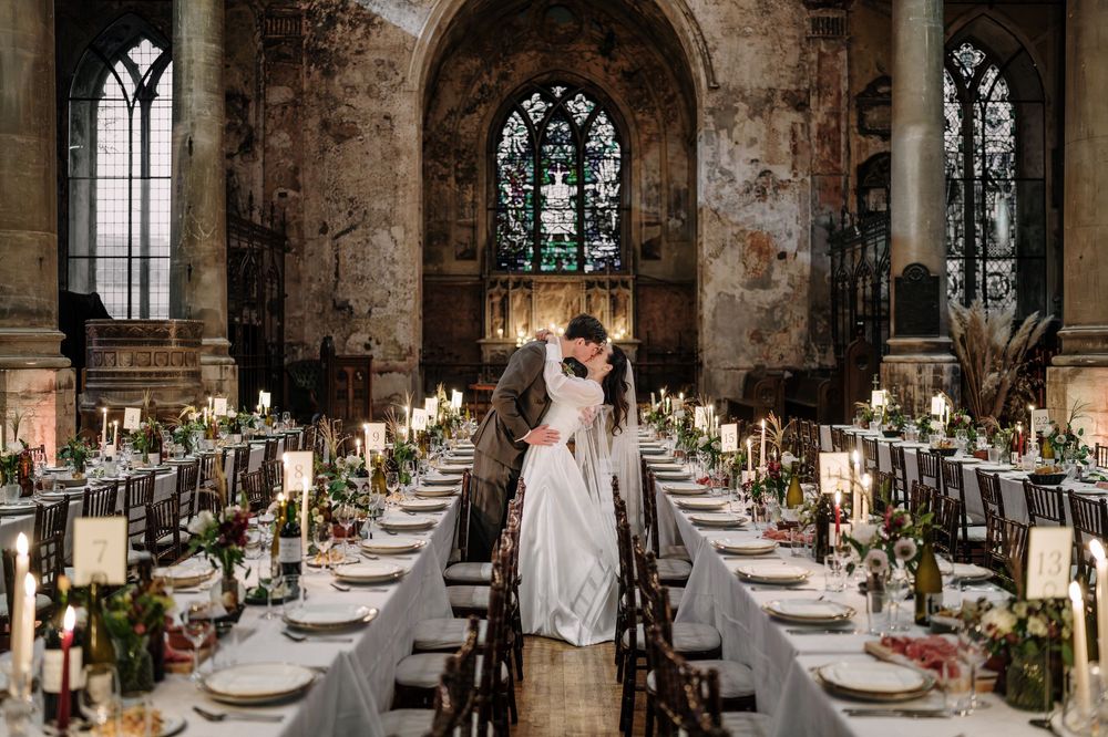Couple kissing at The Mount without Bristol reception with long tables, candles, floral decor, and stunning stained glass windows.