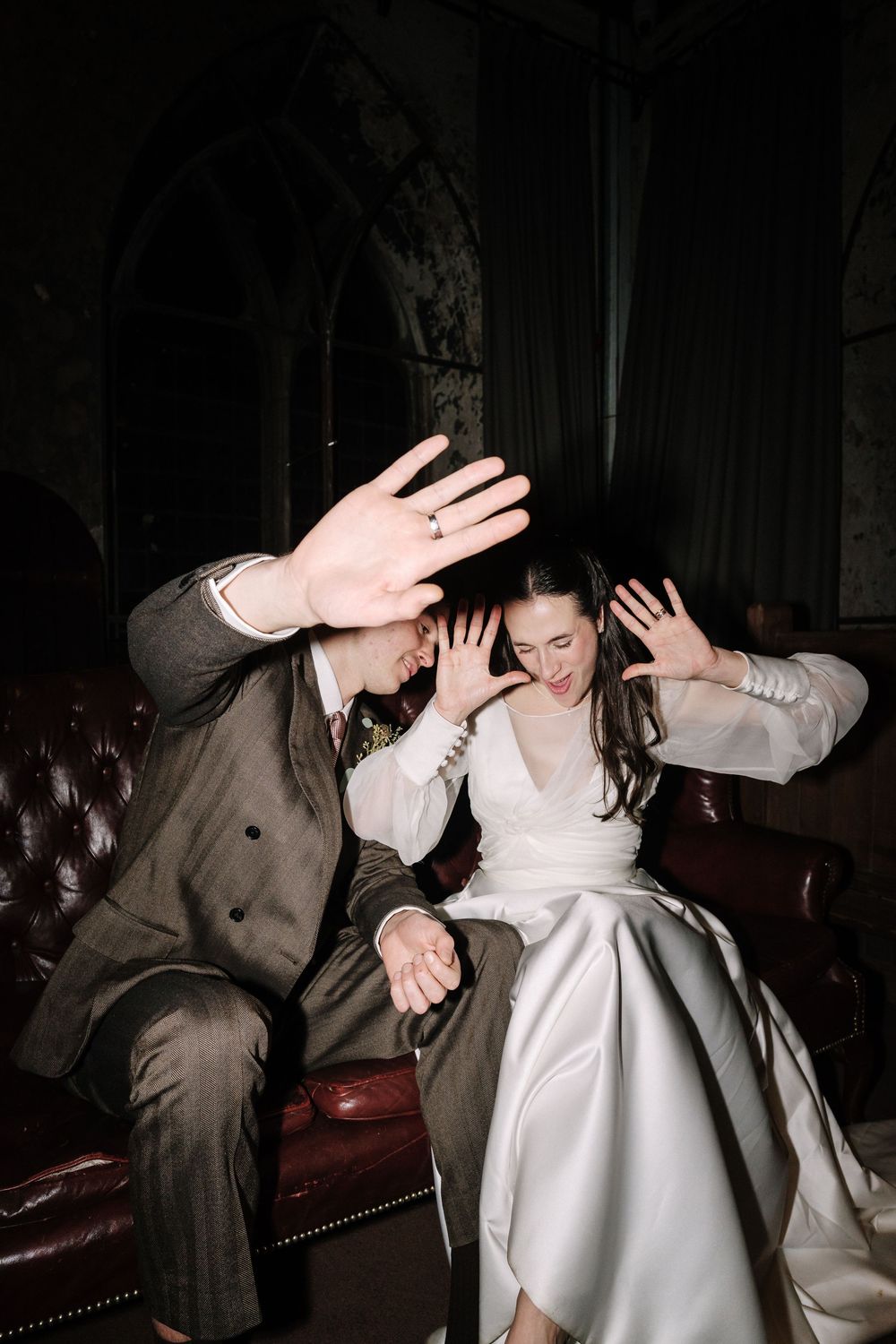 Bride and groom laughing and waving hands playfully on leather sofa at The Mount without Bristol