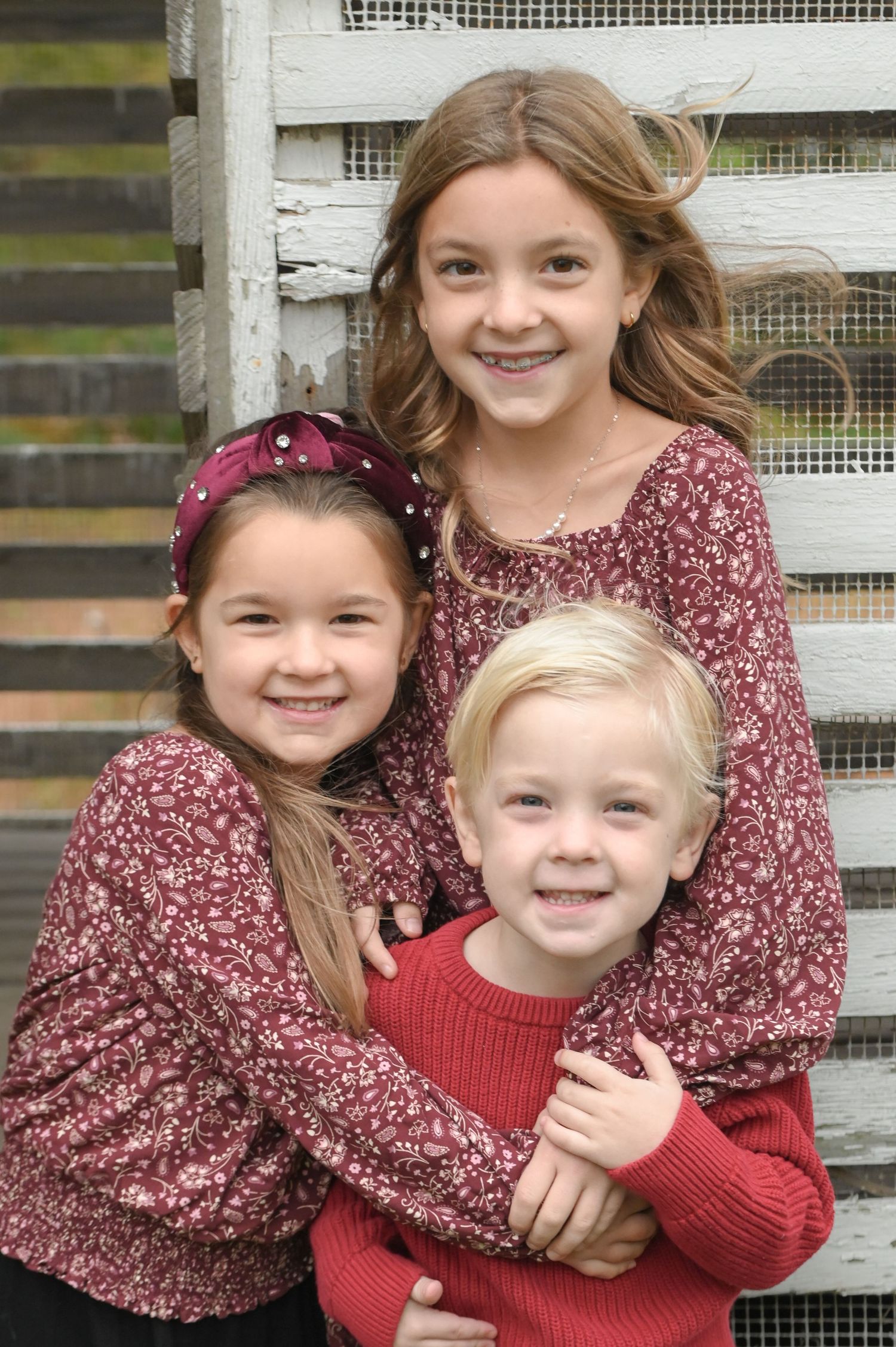 Three smiling children in matching burgundy and red outfits hugging each other in front of a white rustic wooden fence.