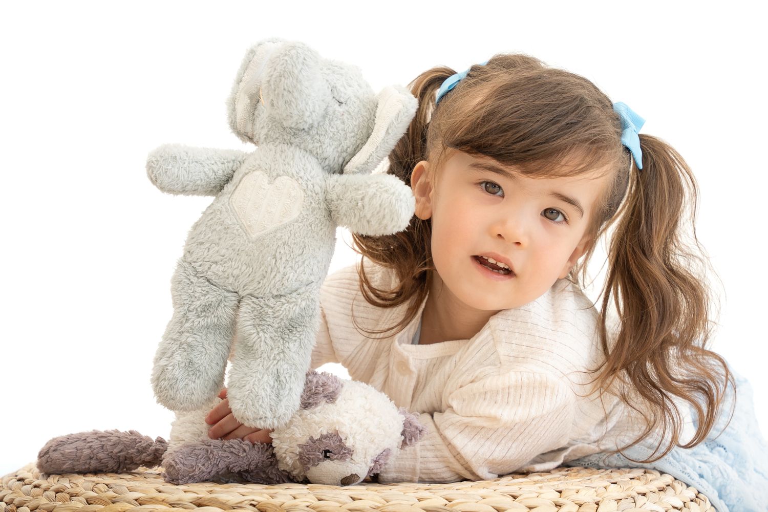 Young child with blue bows in her hair holding a soft grey elephant beside her face, leaning on a woven stool against a cream background.