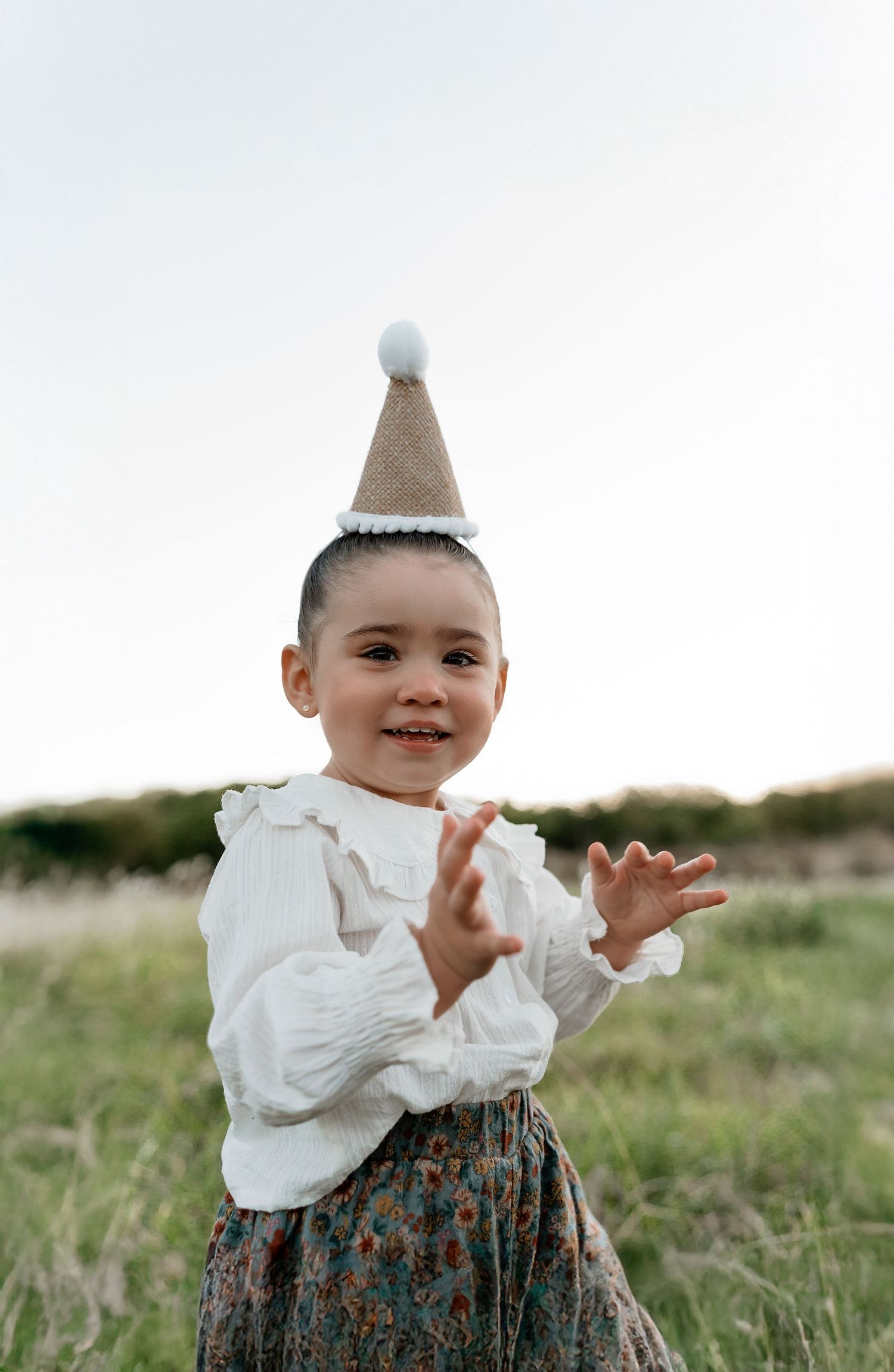 A young child wearing a white ruffled blouse and party hat smiles while playing in a grassy field.