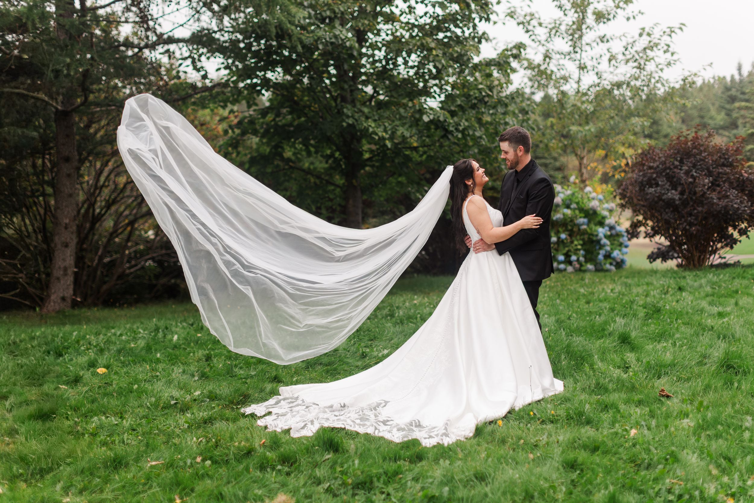 Bride in a white gown with a long sheer veil flowing in the air embraces her groom in a black suit on a lush green lawn at their wedding.