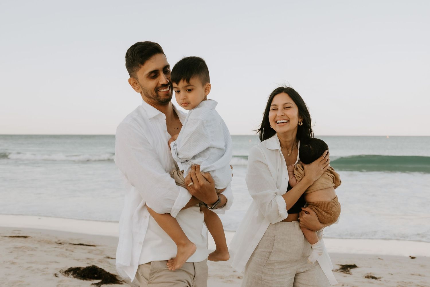 A smiling couple in white linen outfits hold their two young children while walking along a sandy beach with ocean waves behind them.