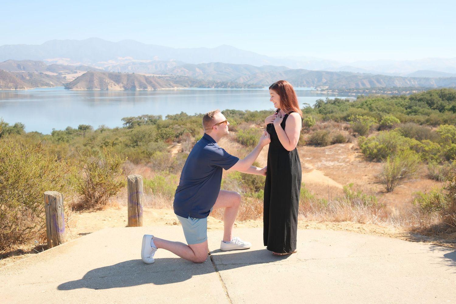 Man proposes on one knee to woman in black dress at scenic lake overlook with mountains in background.