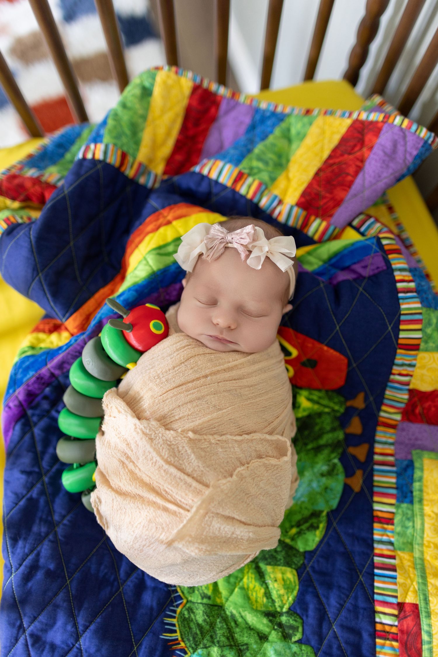 Newborn baby girl swaddled in cream wrap with pink bow headband sleeping in crib on colorful quilt.