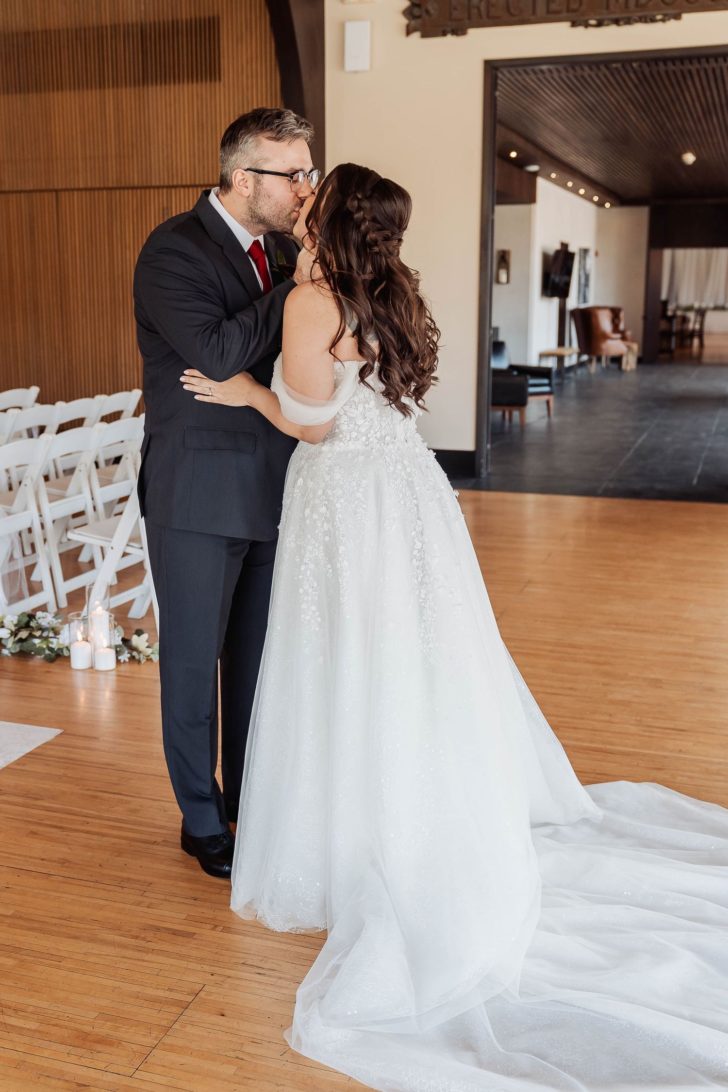 A bride and groom share their first kiss in an elegant indoor wedding venue with white chairs and hardwood floors.