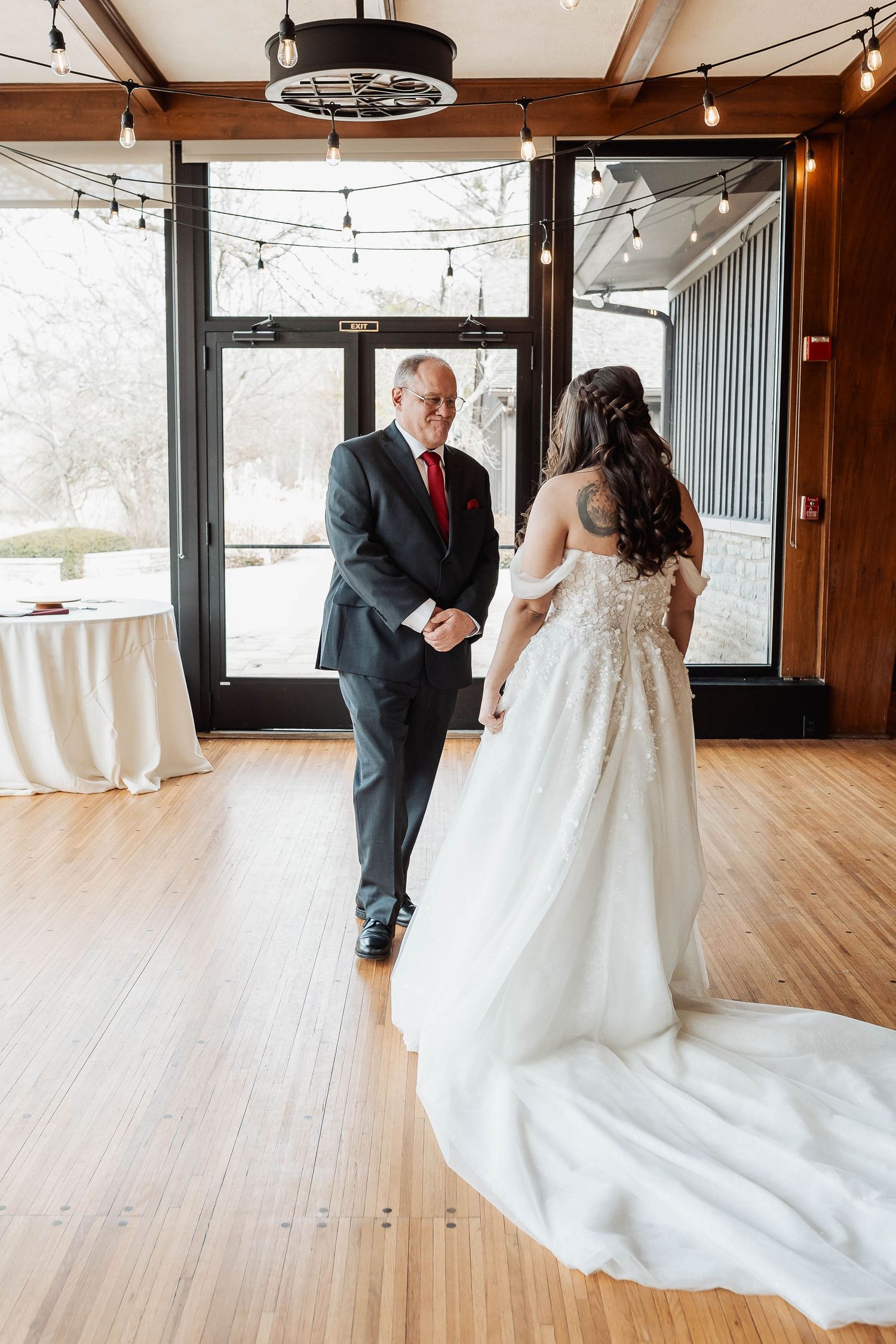 Bride and father share first look moment in elegant wedding venue with string lights and hardwood floors.