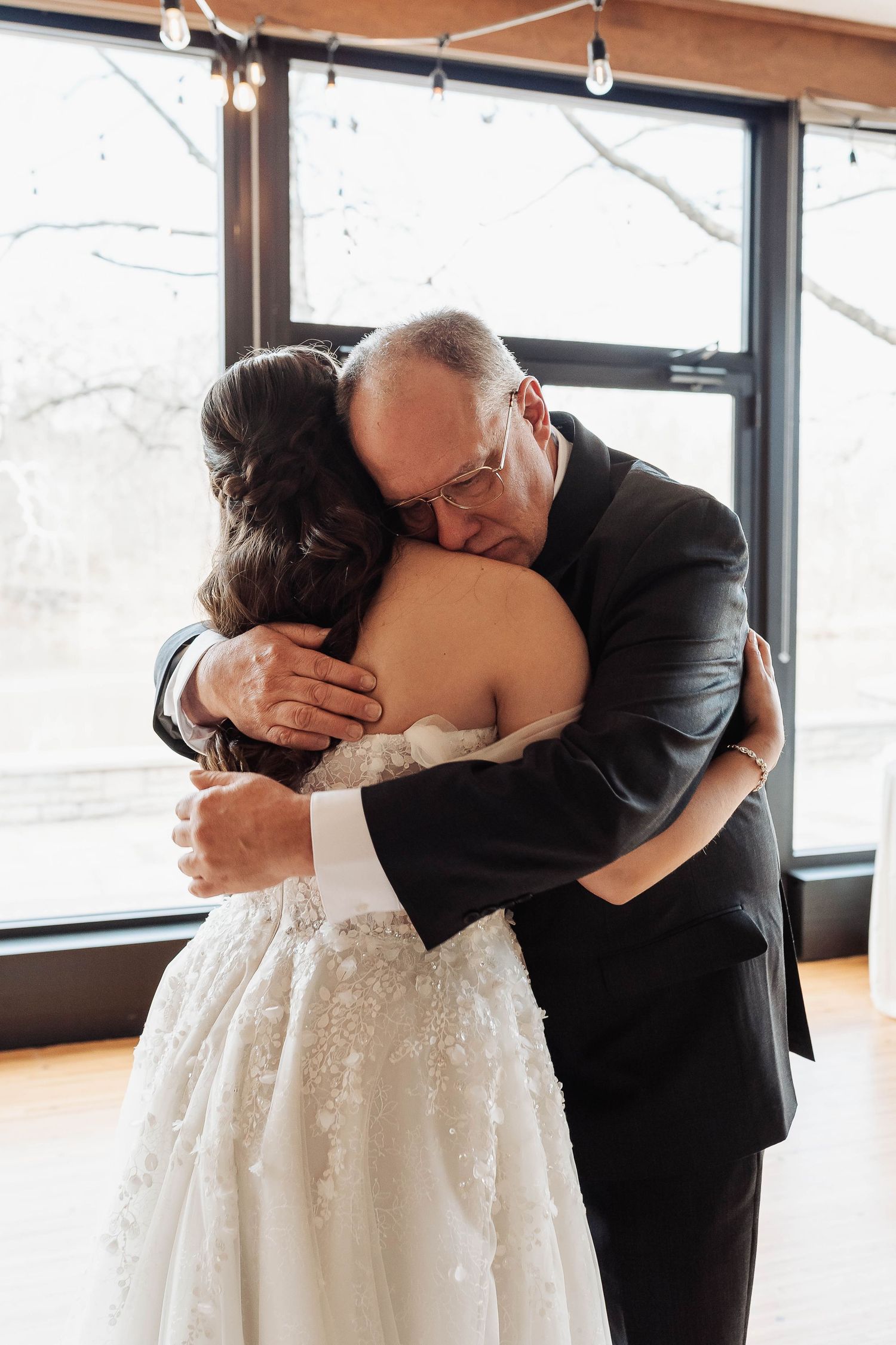 Bride in lace wedding gown shares emotional hug with father in black suit during wedding reception.