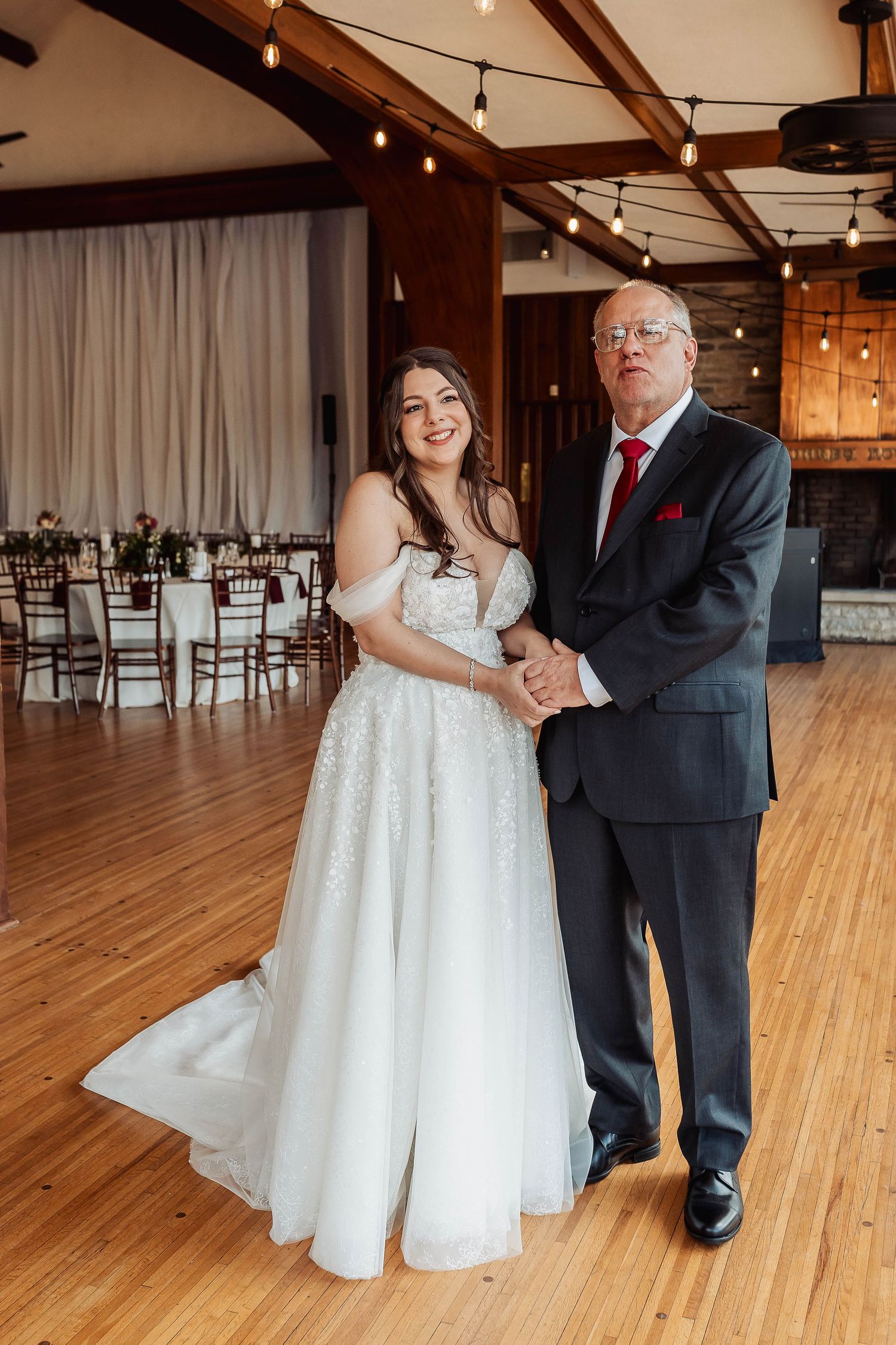 Bride in white gown stands with older gentleman in dark suit and red tie in elegant wedding reception venue.