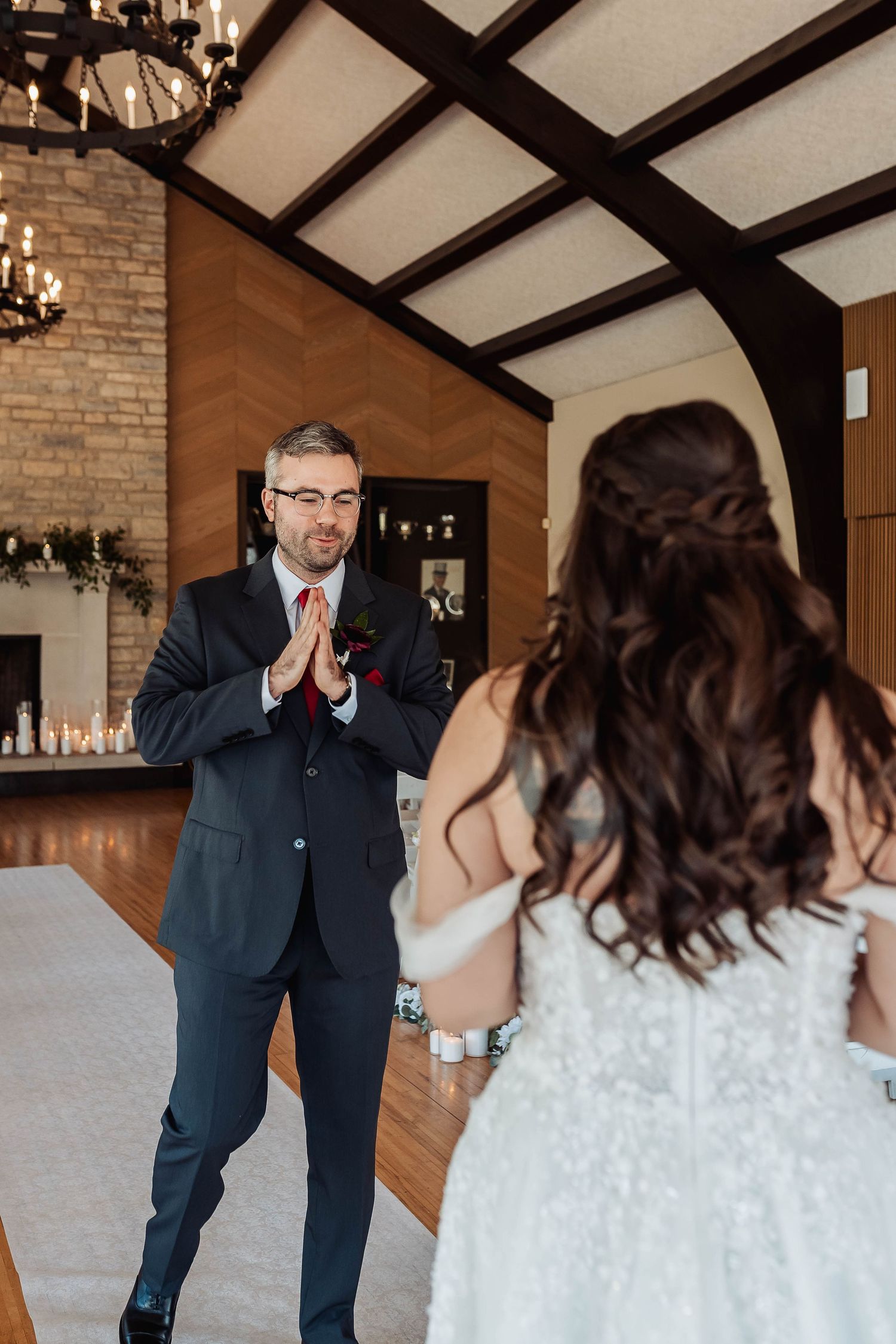 Groom in dark suit clasps hands in awe seeing bride in white wedding dress during first look ceremony indoors.