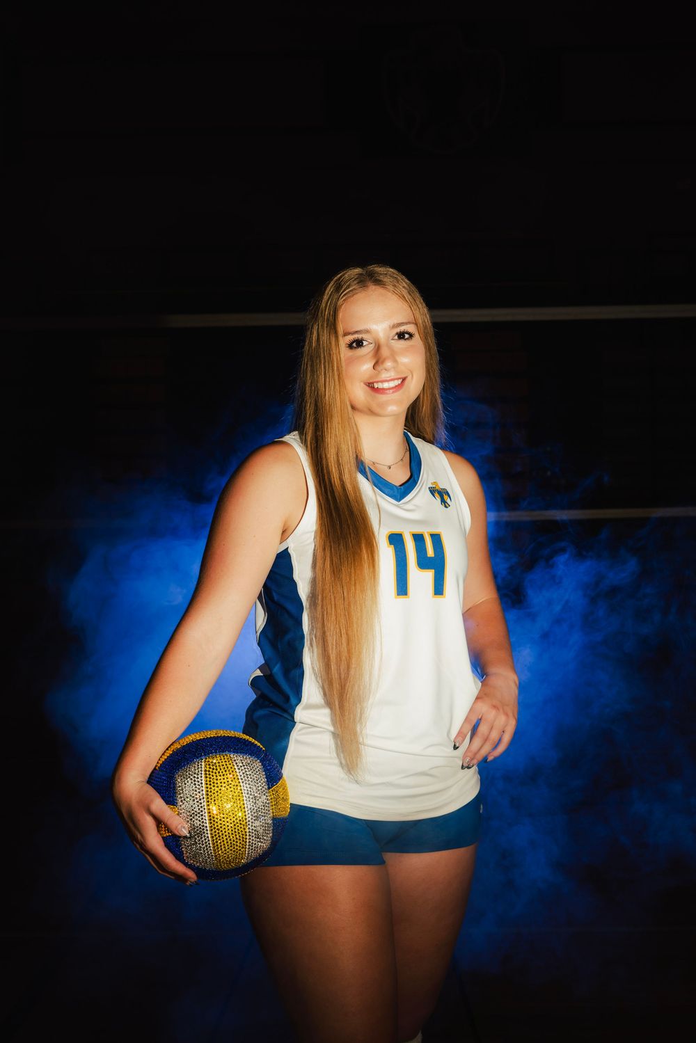 Female volleyball player #14 in white and blue uniform holding a ball, posed against dramatic blue smoke background.