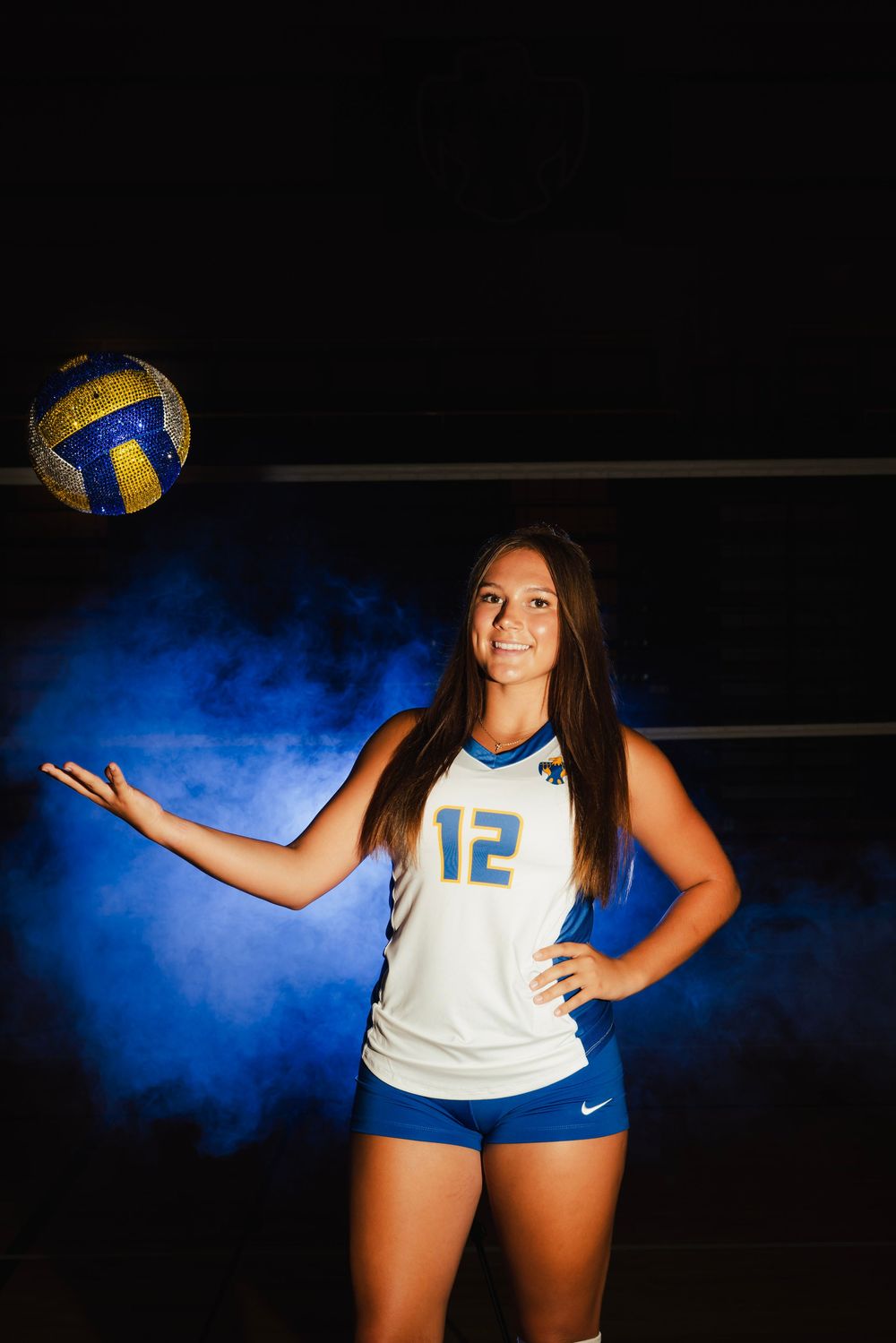 Female volleyball player #12 in blue and white Nike uniform poses with ball in dramatic blue smoke lighting.