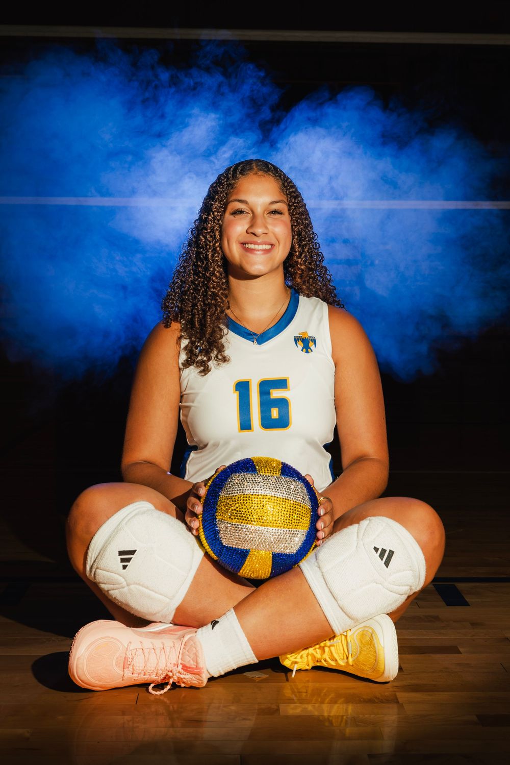 Volleyball player #16 sits cross-legged on court holding ball, wearing white uniform, with blue smoke background.