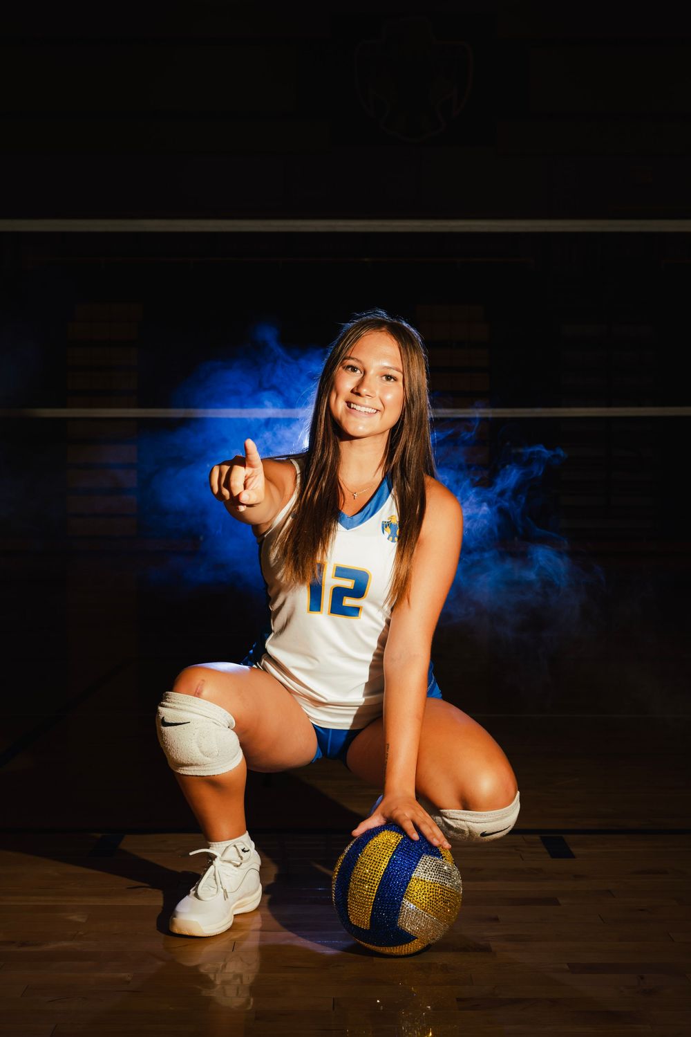 Female volleyball player #12 kneeling with ball, pointing at camera in dramatic blue smoke lighting.