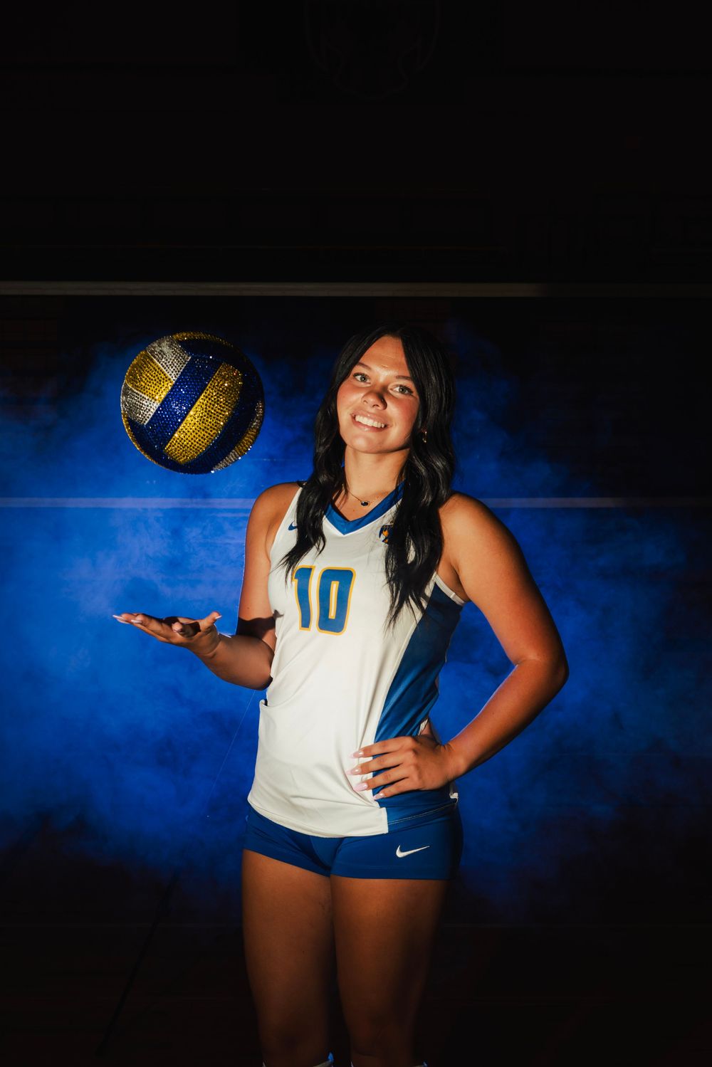 Female volleyball player #10 in blue and white Nike uniform tosses a glittery ball against a dramatic blue smoky background.