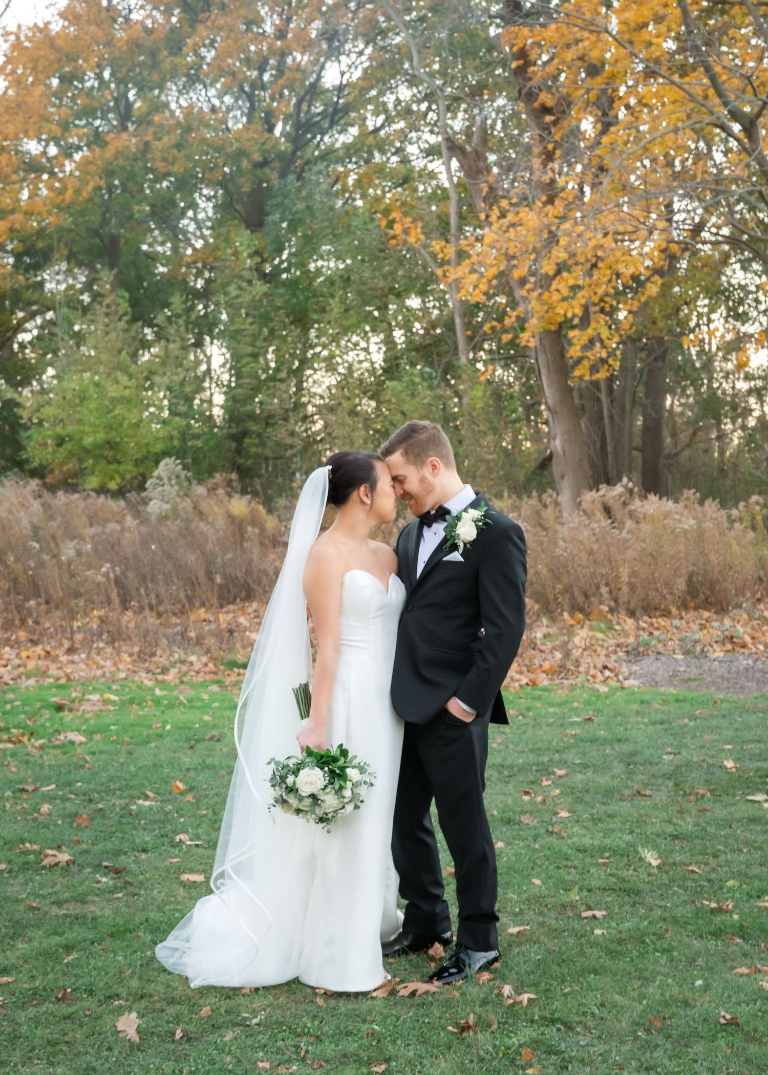 Bride in a white gown and veil and groom in a black suit embrace foreheads together on a grassy lawn surrounded by autumn trees.