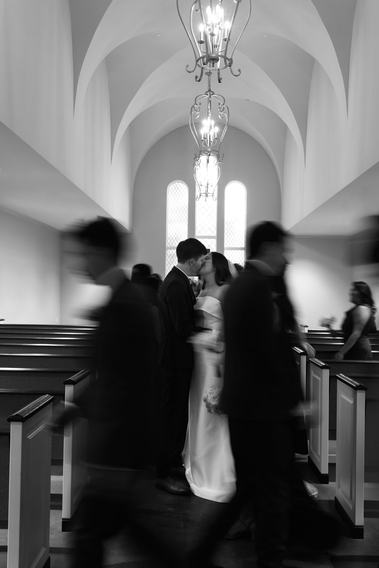 Black and white photo of a couple kissing at the altar in a church aisle during their wedding ceremony.