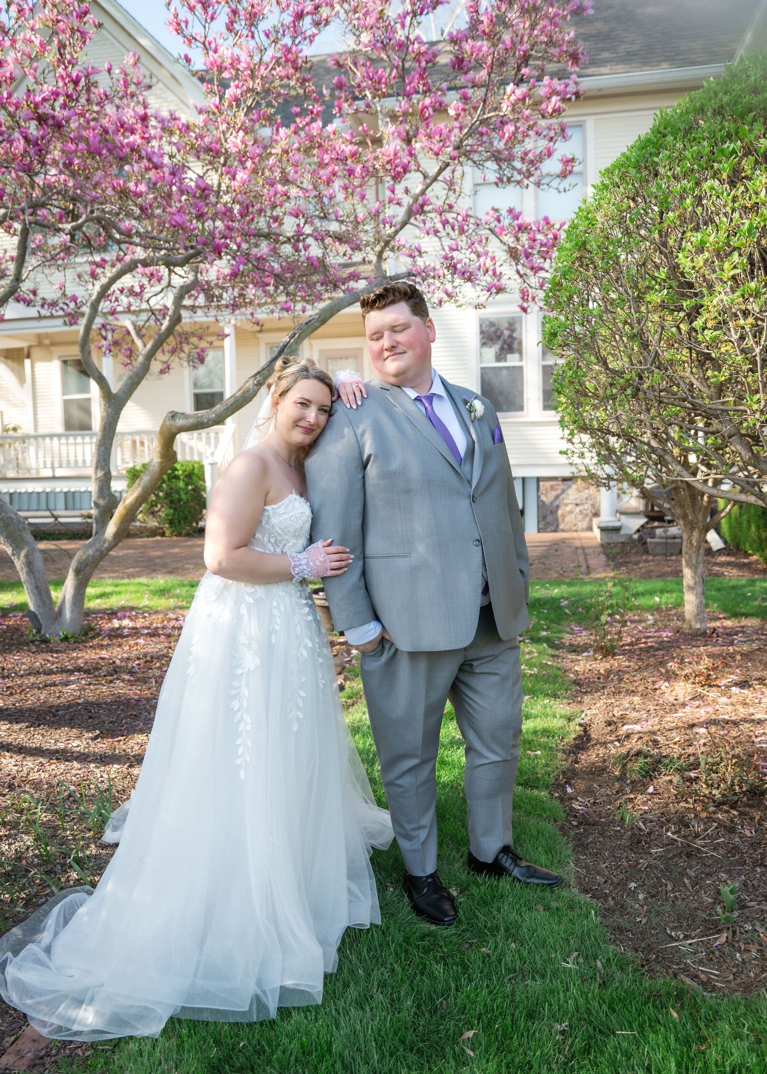 Smiling bride in a white lace gown and groom in a gray suit with purple tie pose together on a lawn beneath a blooming magnolia tree.