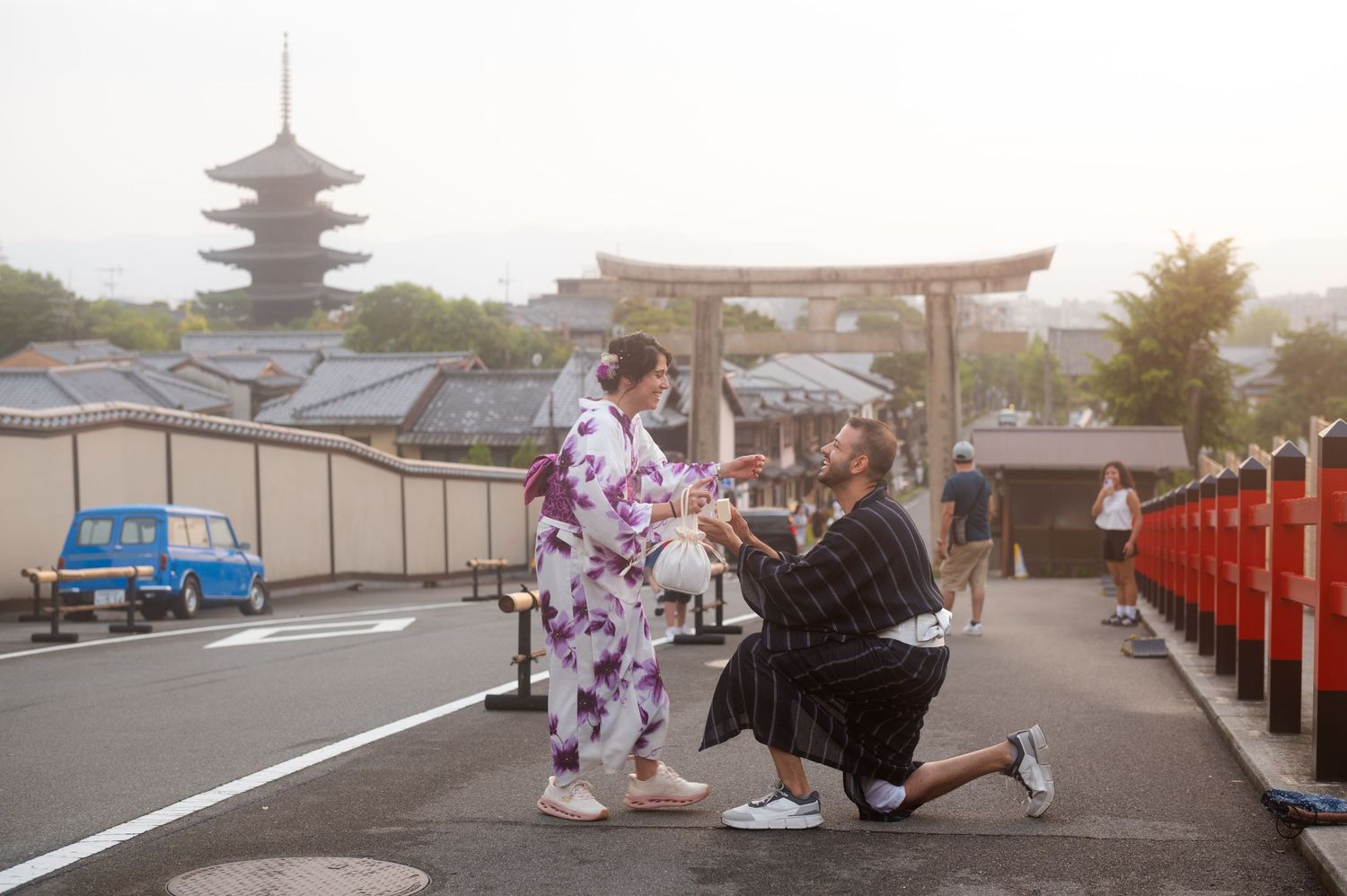 Man in a dark kimono kneels on one knee proposing to a woman in a floral kimono near a Japanese torii gate and pagoda.