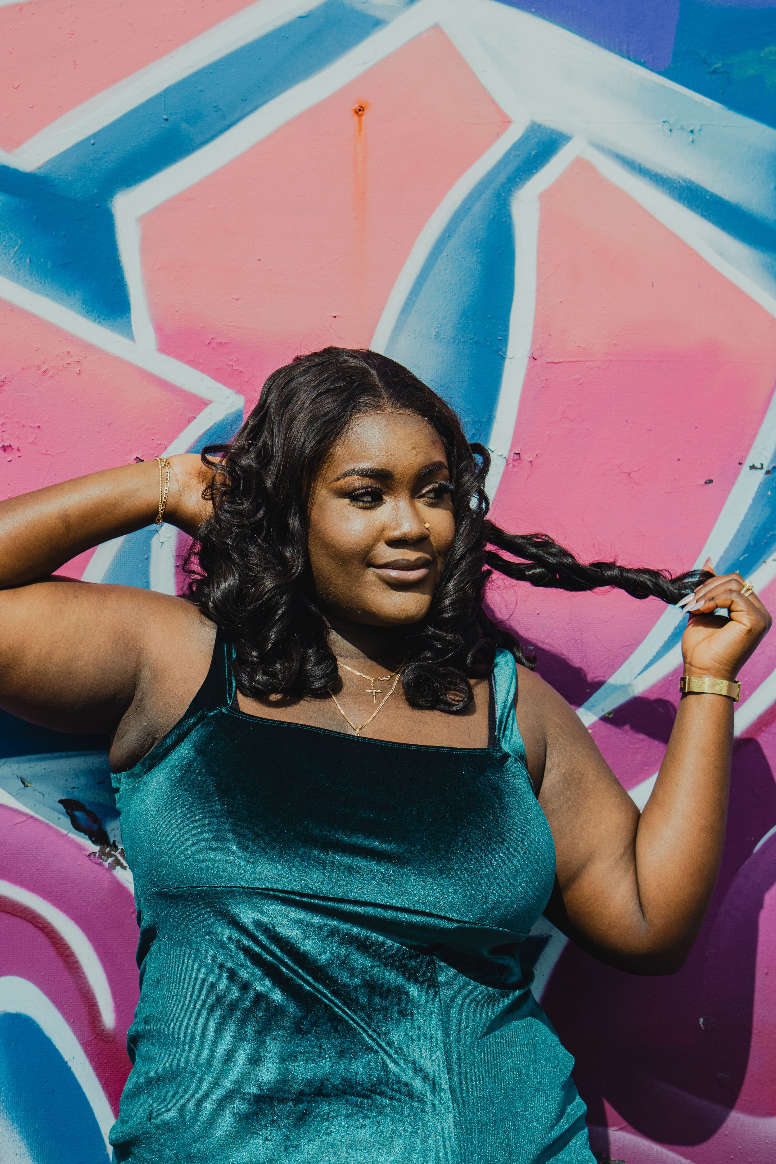 Woman in teal velvet dress poses confidently against colorful graffiti wall, styling her long dark hair.