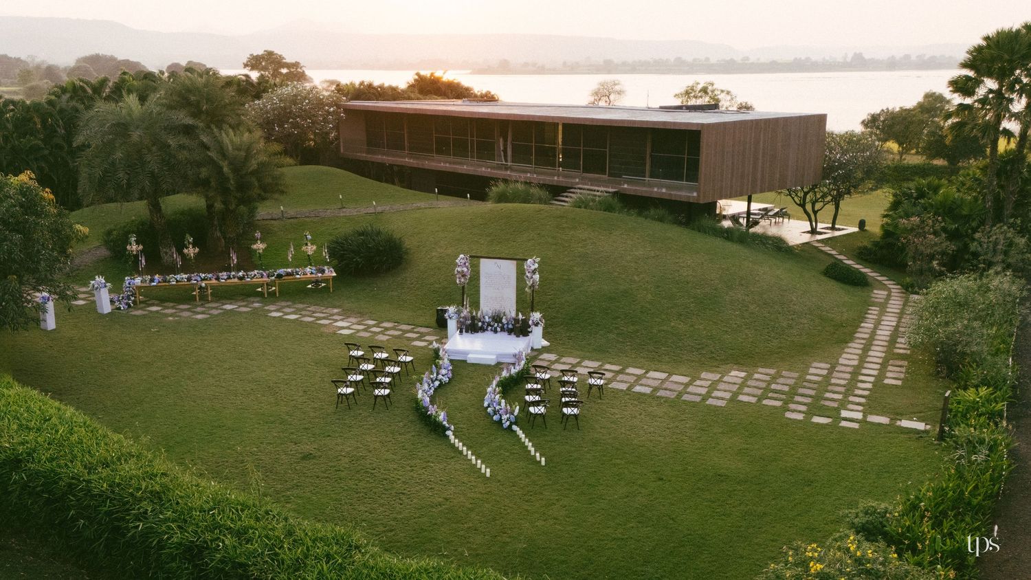 Aerial view of an outdoor garden wedding ceremony with a floral aisle, decorated altar, candlelit tables and a modern wooden venue.