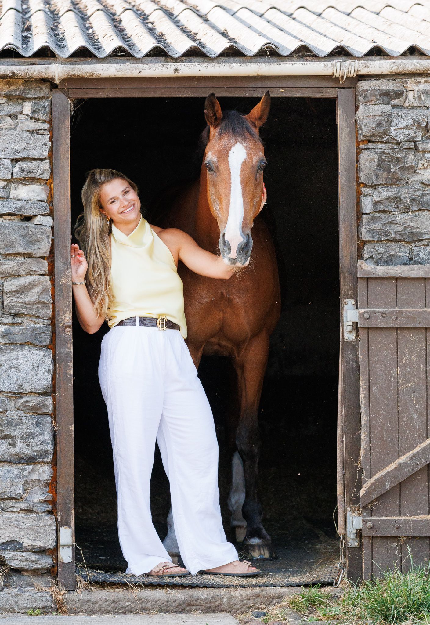Young woman smiling between two bay horses with white markings, holding reins near a brick stable.