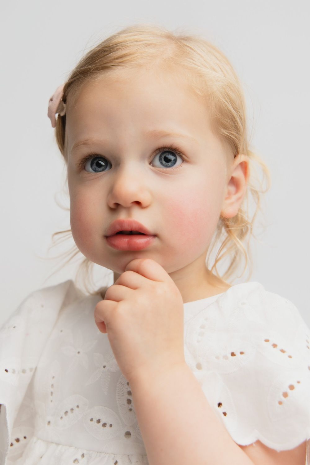 Adorable blonde toddler girl with blue eyes wearing a white eyelet dress, looking curiously with hand near chin.