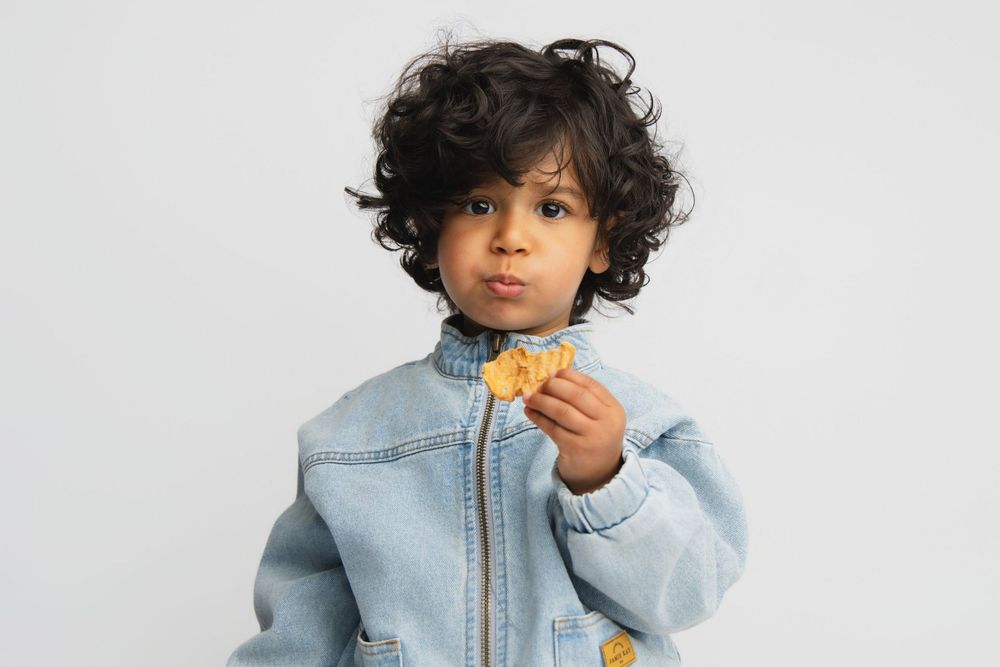 Young child with curly dark hair wearing a denim jacket, holding a small snack against a white background.