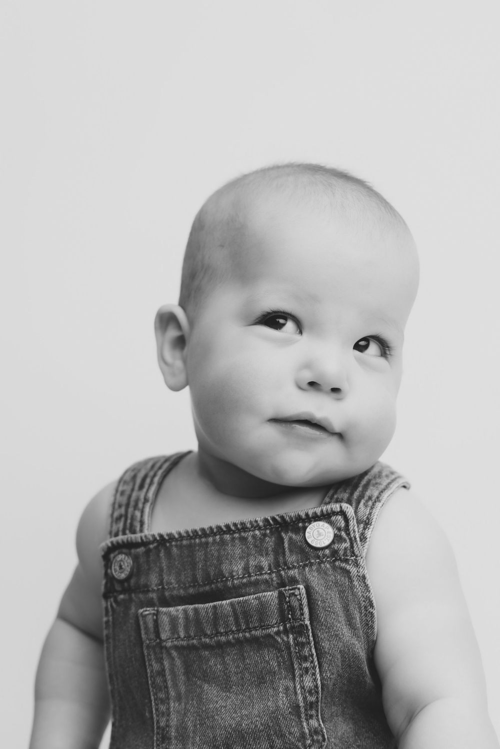 Black and white portrait of an adorable baby in denim overalls looking up curiously against a light background.