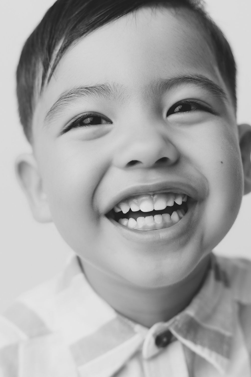 Black and white photo of a joyful young Asian boy laughing in a plaid shirt.