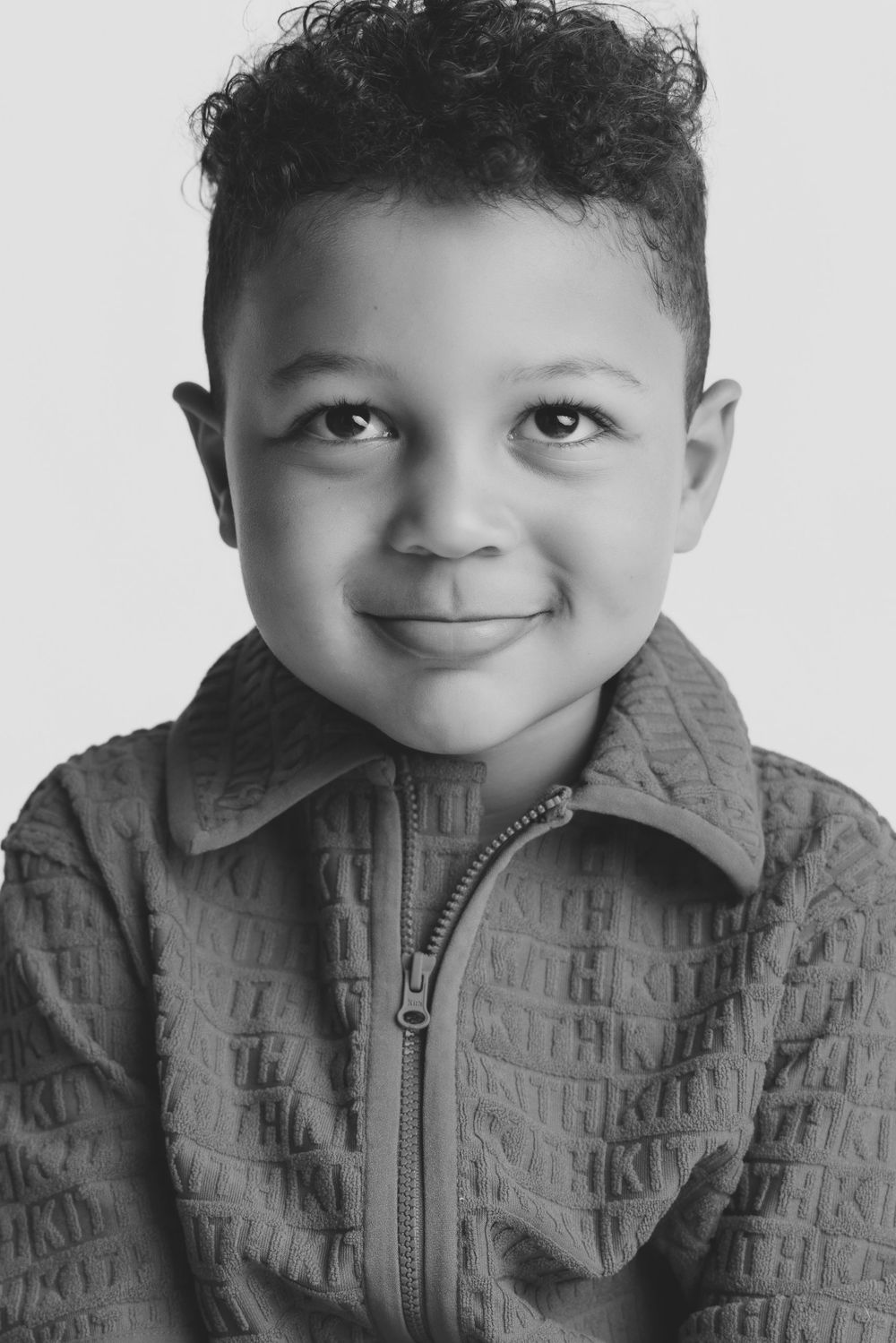 Black and white portrait of a smiling young boy with curly hair wearing a zip-up jacket.