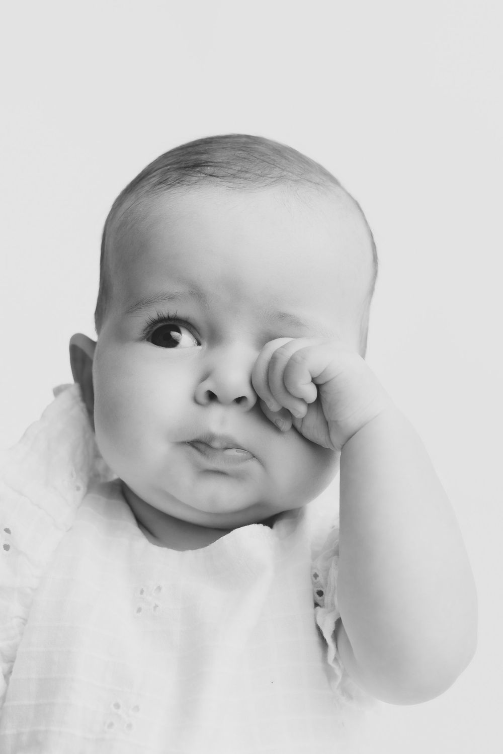 Black and white portrait of a sleepy baby rubbing its eye with a tiny fist, wearing a white outfit.